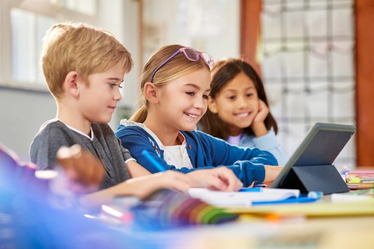 Happy multiethnic elementary classmates working on digital tablet in classroom during lesson. Three elementary school pupils using digital tablet at desk during technology class. Schoolchildren in class using technology for studying together in a interactive lesson.